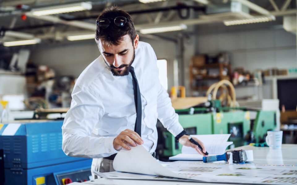 Young hardworking caucasian bearded controller looking at printed sheets and evaluates quality while standing in printing shop. Young hardworking caucasian bearded controller looking at printed sheets and evaluates quality while standing in printing shop.